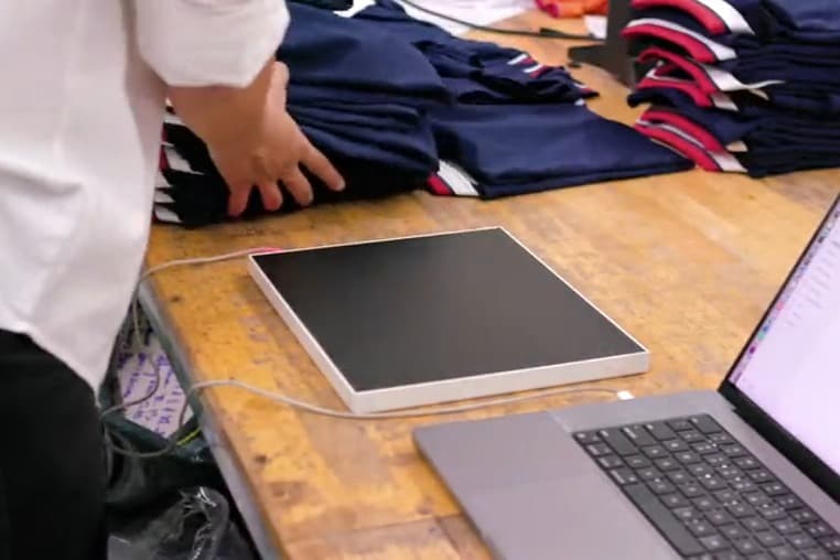 Đầu đọc RFID để bàn nối với laptop, đặt ngay cạnh chồng áo đã gấp trên bàn soạn hàng